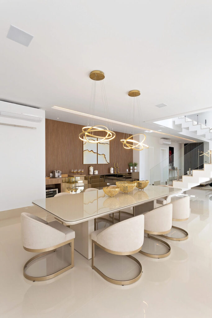 Sala de jantar com mesa retangular de vidro e cadeiras estofadas em tom creme, com base dourada. Acima, duas luminárias modernas de formato circular, com acabamento dourado e luz quente amarela, iluminam a mesa de forma sofisticada. Ao fundo, uma parede com ripas de madeira e um quadro abstrato complementam a decoração elegante. O ambiente exibe um estilo contemporâneo e minimalista, ideal para recepções e jantares sofisticados