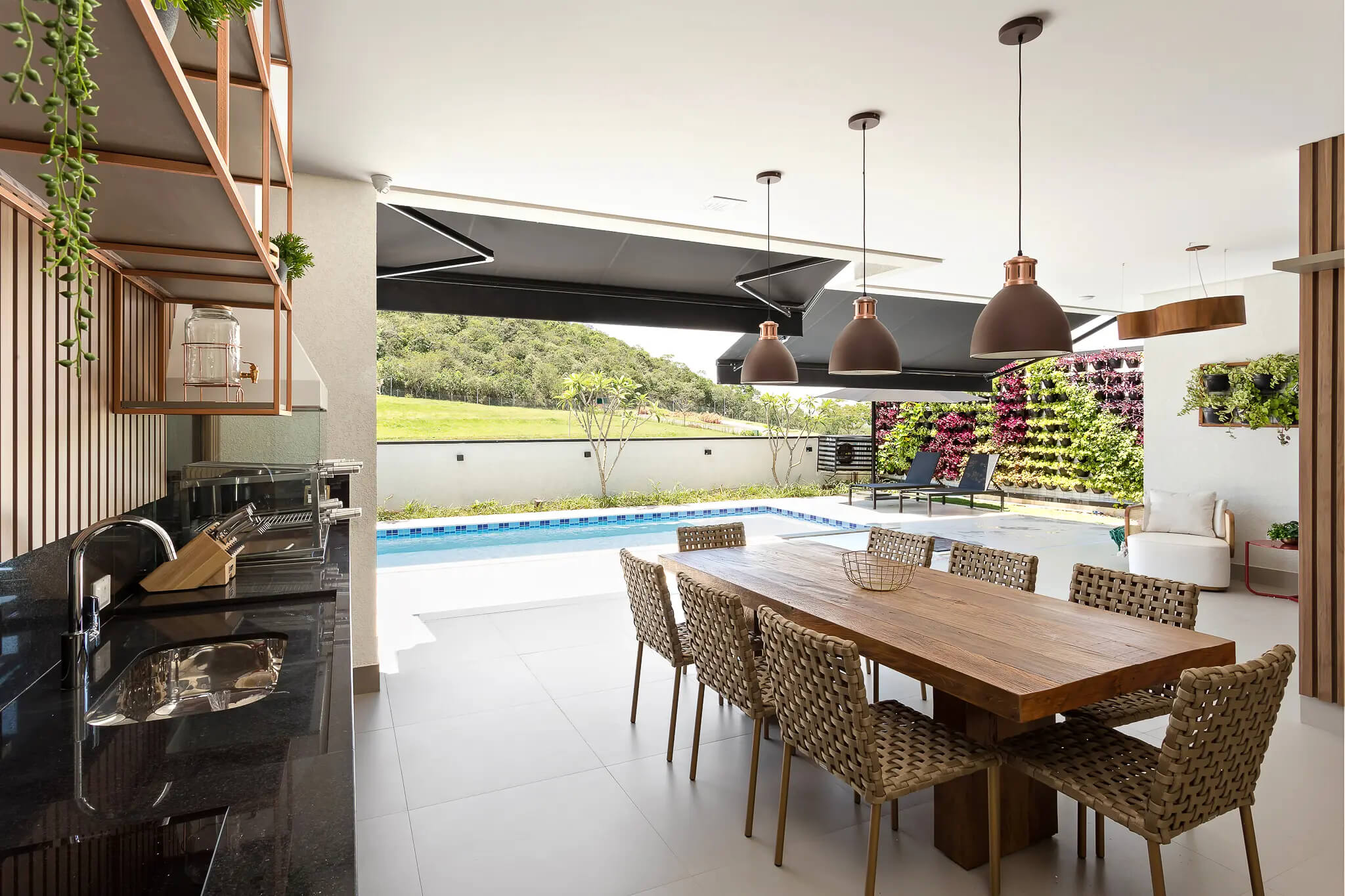 Espaço gourmet com linda e grande mesa de madeira, cadeiras ao redor e piscina visível ao fundo, destacando a importância da iluminação na decoração, com pendentes visíveis realçando o ambiente