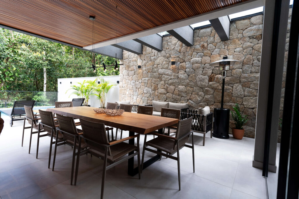 Espaço gourmet com parede de pedra, teto de madeira e uma grande mesa de madeira com cadeiras ao redor, destacando o uso de diferentes elementos de iluminação, como spots, para realçar a decoração de forma elegante