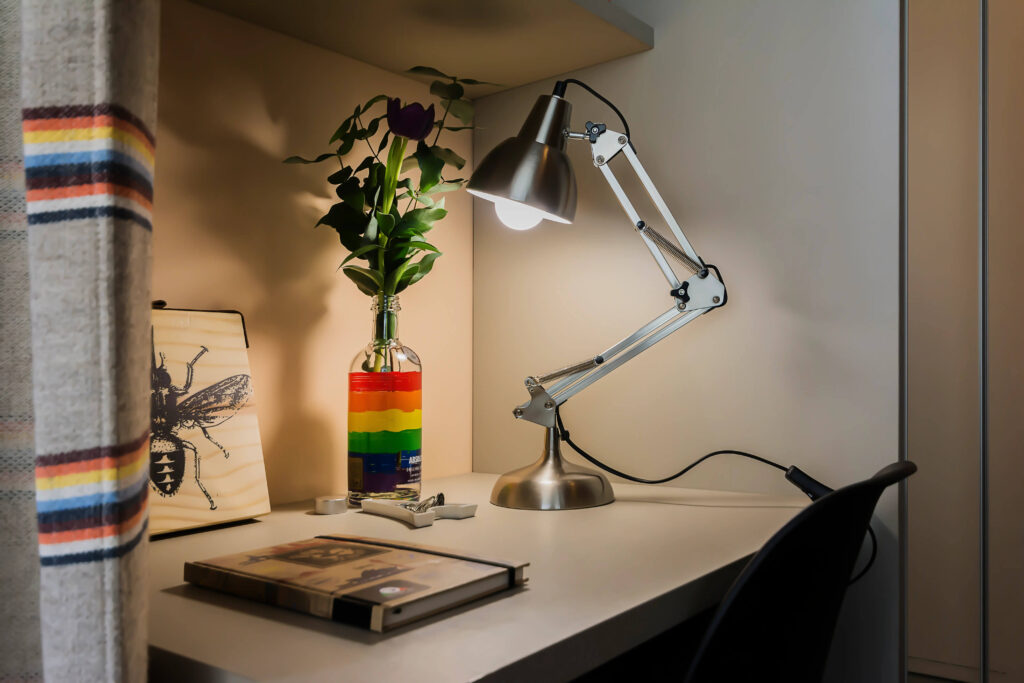 Um cantinho acolhedor e vibrante para leitura, com uma luminária direcionável iluminando uma mesa simples. Ao fundo, uma garrafa colorida em tons do arco-íris com flores frescas, criando uma atmosfera alegre e convidativa, ideal para momentos de relaxamento e leitura.