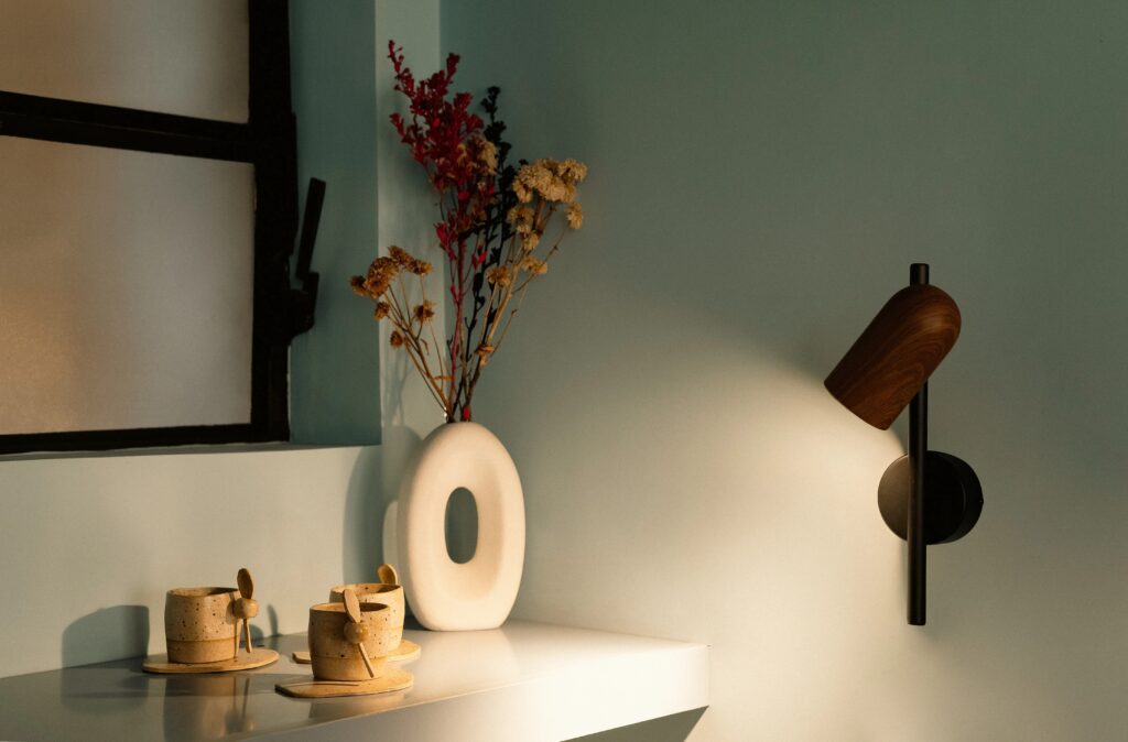 Um ambiente minimalista e sereno, com iluminação suave, apresenta uma luminária de parede moderna, flores secas em um vaso branco e duas xícaras de cerâmica sobre uma mesa branca.