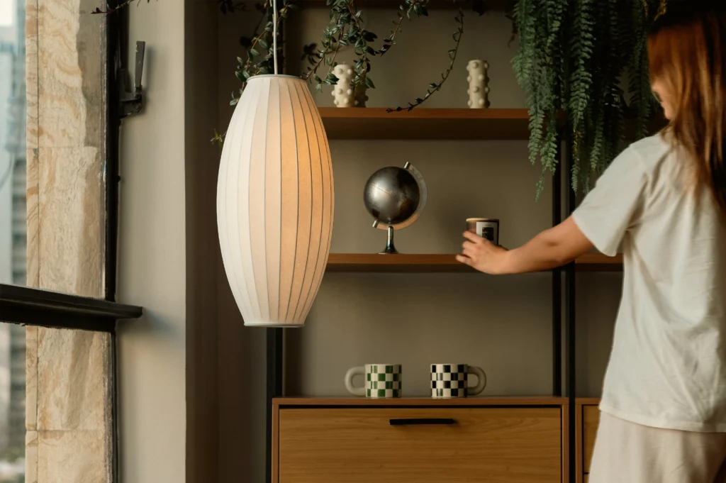 Uma pessoa com uma camisa clara ajusta um objeto em prateleiras de madeira que exibem um pequeno globo e canecas. Uma luminária pendente moderna emite um brilho suave, com plantas suspensas ao redor. A cena transmite aconchego e organização.
