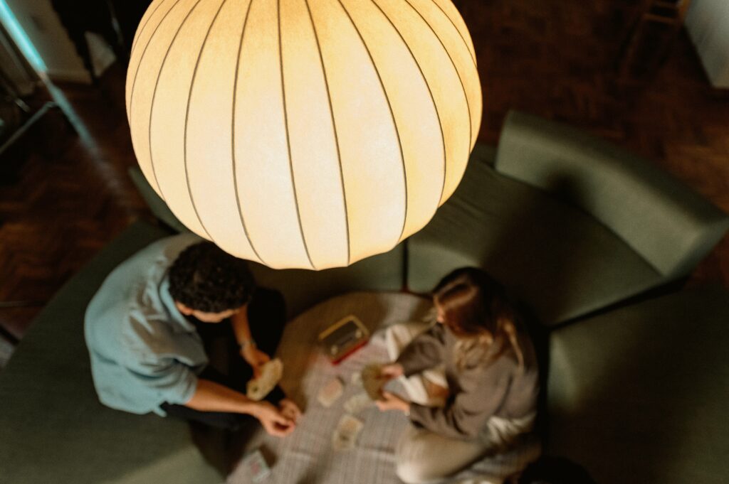 Duas pessoas sentadas em um sofá verde jogam um jogo de cartas sobre um cobertor, sob uma grande luminária pendente de luz quente. A cena é aconchegante e intimista.