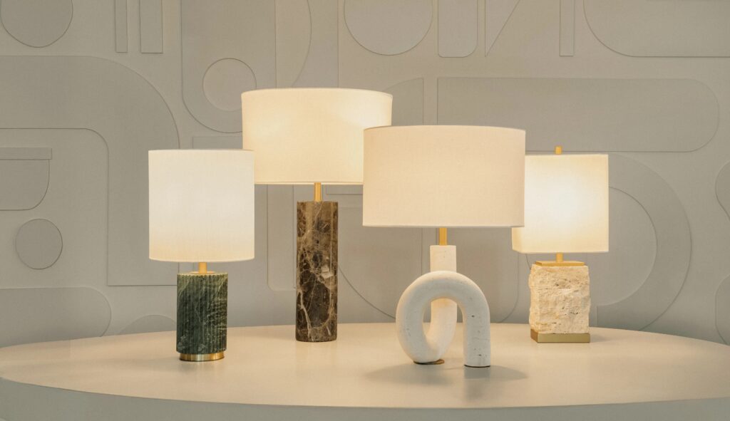 Um conjunto de quatro luminárias de mesa sobre uma superfície branca curvada, cada uma com bases texturizadas e únicas em materiais como mármore e pedra, posicionadas diante de uma parede estampada.