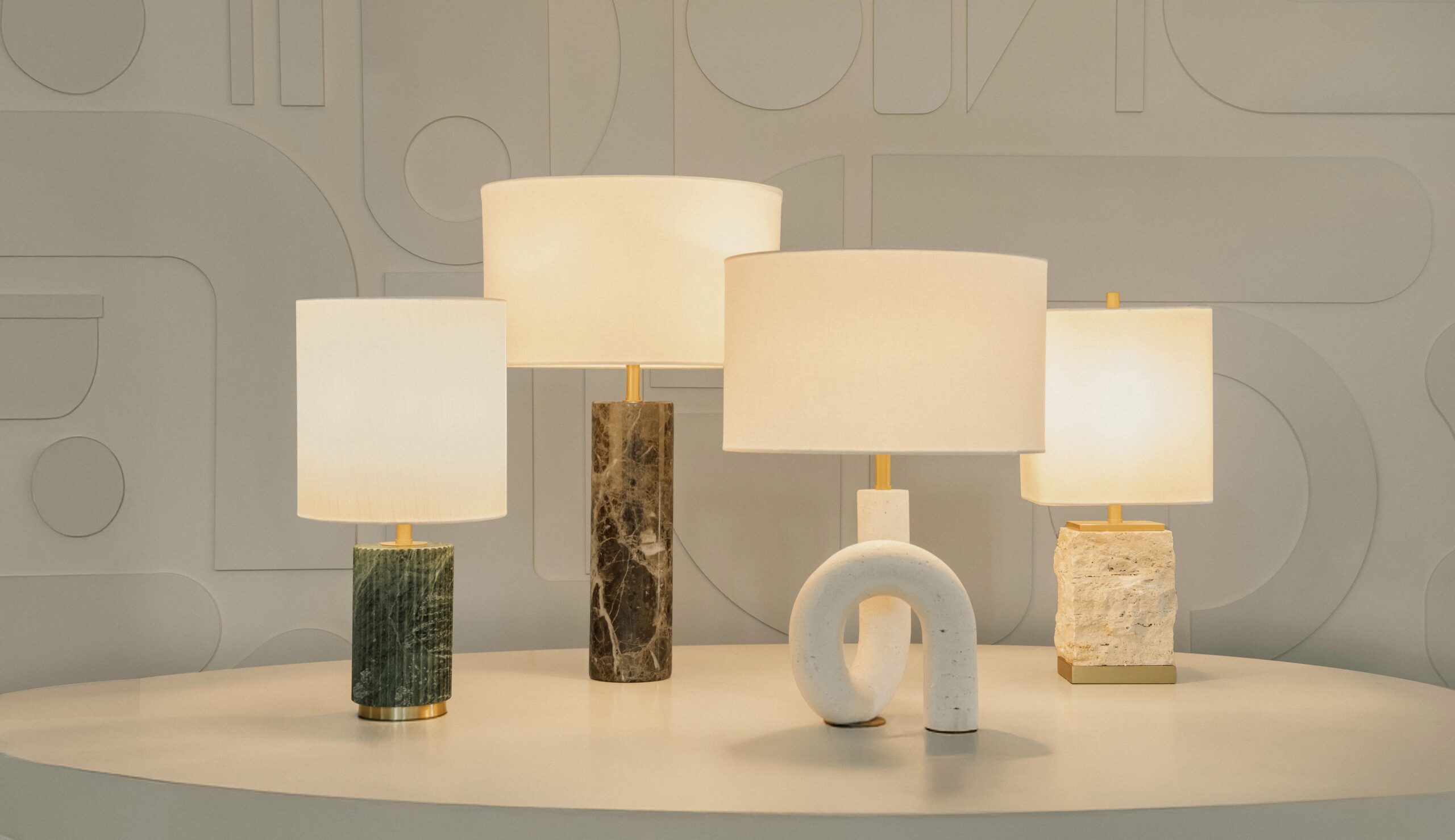 Um conjunto de quatro luminárias de mesa sobre uma superfície branca curvada, cada uma com bases texturizadas e únicas em materiais como mármore e pedra, posicionadas diante de uma parede estampada.