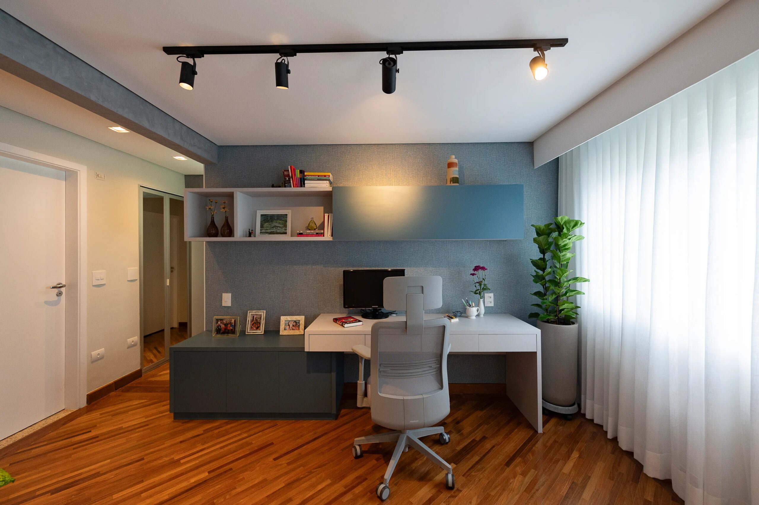 Home office bem iluminado com mesa de trabalho branca, cadeira ergonômica, estantes de parede com livros e objetos decorativos, planta grande ao lado da janela de cortinas brancas, piso de madeira e iluminação direcionada no teto.