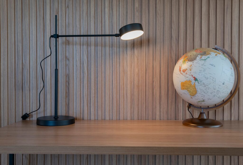 Luminária de mesa moderna acesa ao lado de um globo terrestre em uma mesa de madeira com fundo de painel de madeira vertical, criando um ambiente de estudo ou trabalho.