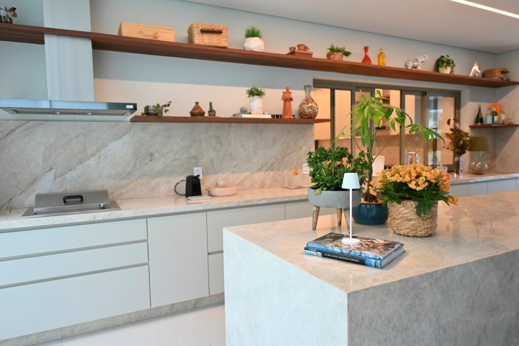 Imagem de uma cozinha moderna decorada com plantas verdes e flores, com balcões de mármore branco, prateleiras de madeira e uma atmosfera acolhedora.