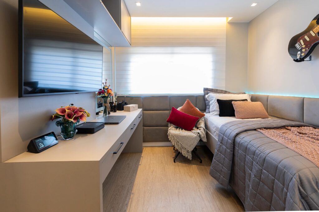 Quarto moderno decorado com televisão de tela plana, almofadas coloridas, guitarra decorativa na parede e bancada com flores e gadgets, iluminação suave.